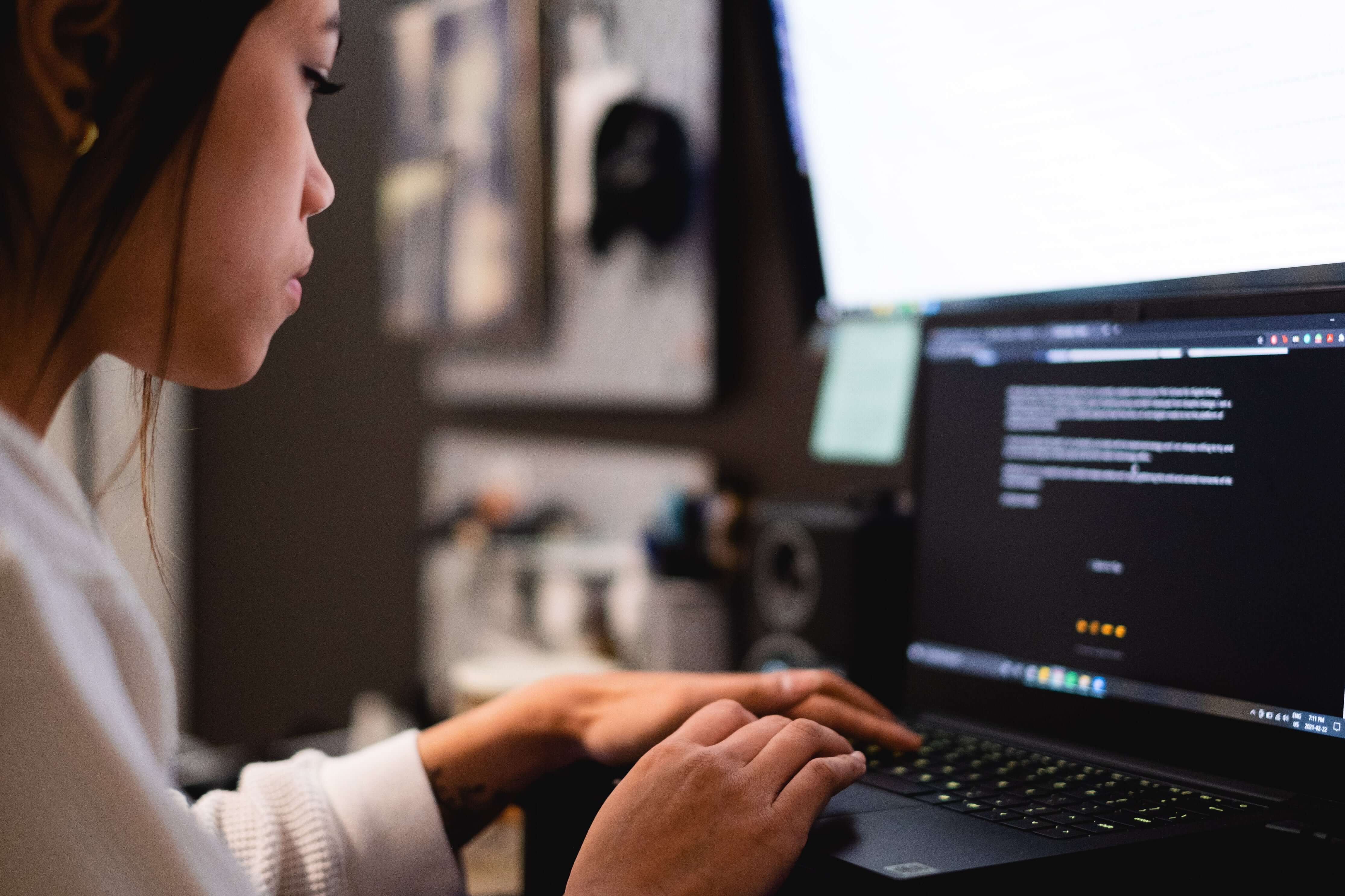 Person typing on a laptop with a computer screen displaying code in a home office setting.
