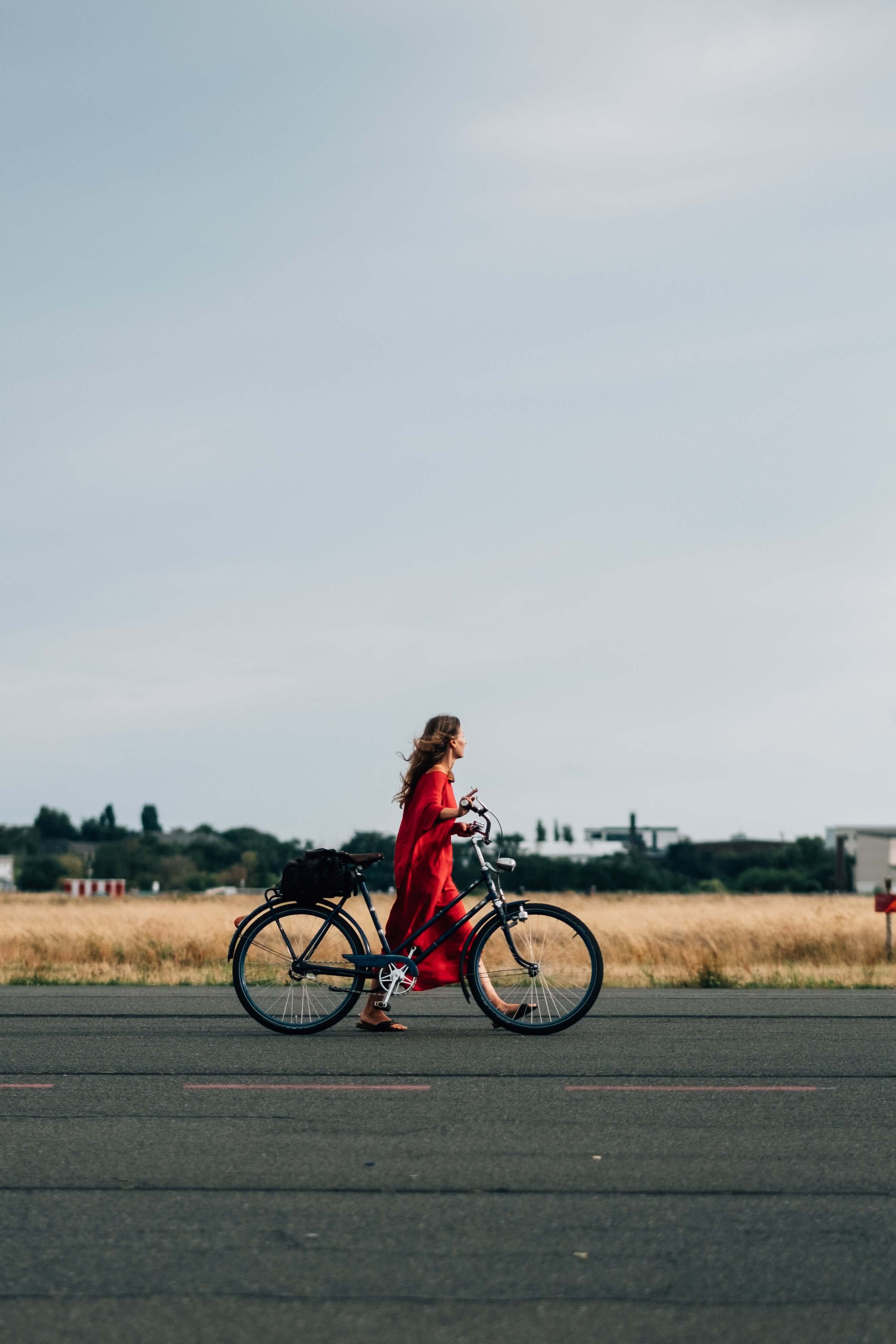 Person in a red dress pushing a bicycle on an open road with a clear sky.
