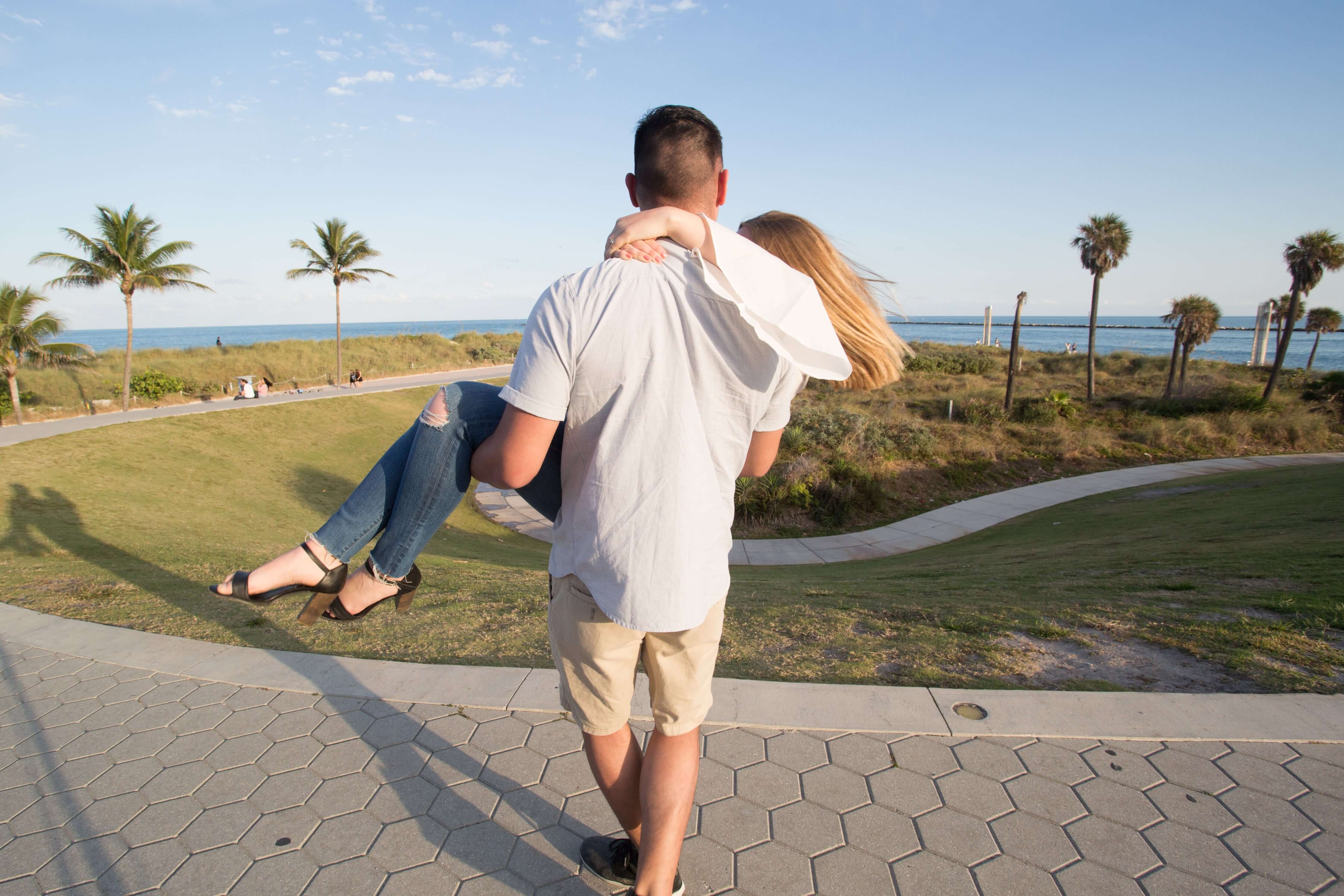 Man lifting a woman on a beach path with palm trees and ocean in the background
