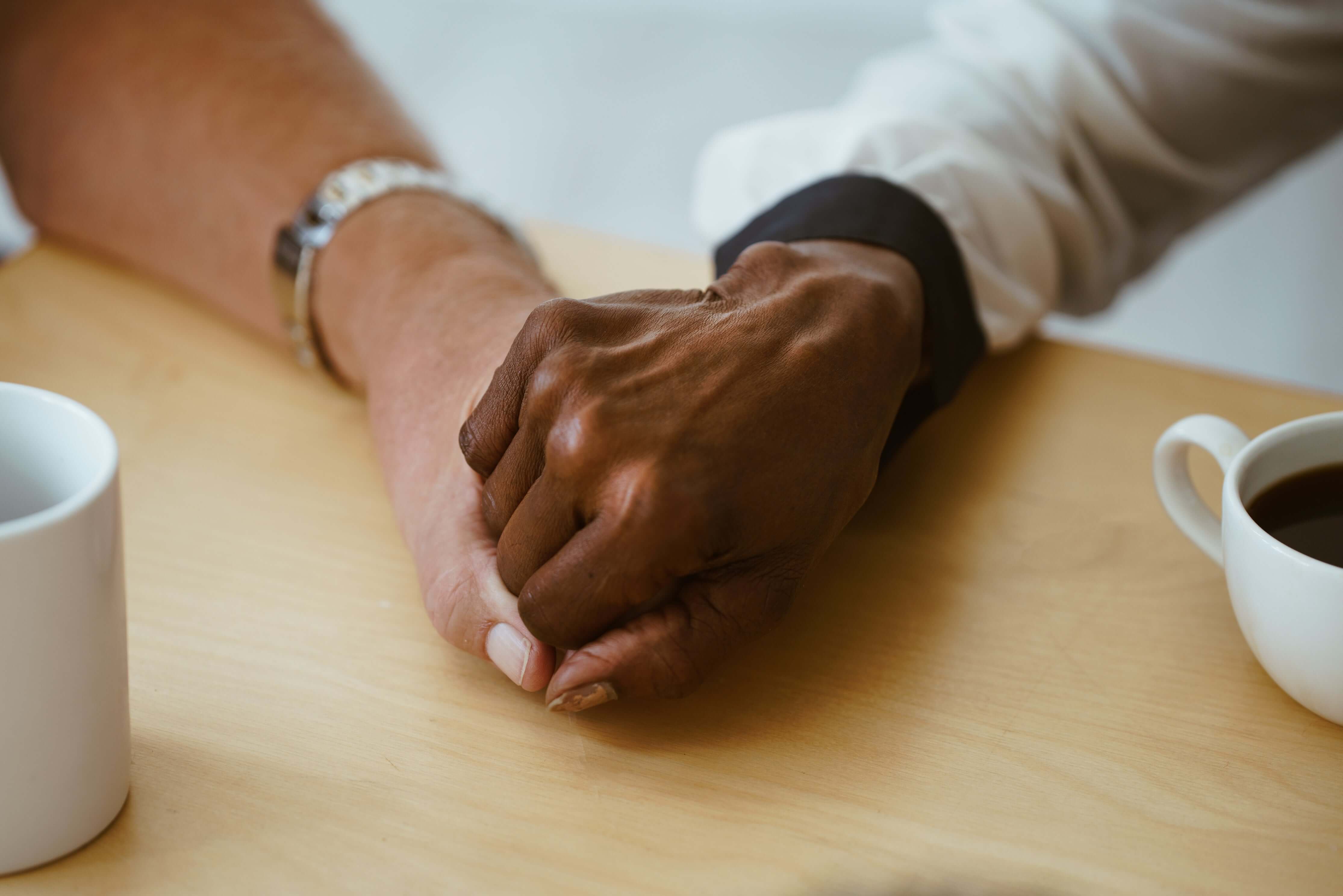 Two people's hands clasped together on a table with coffee cups.
