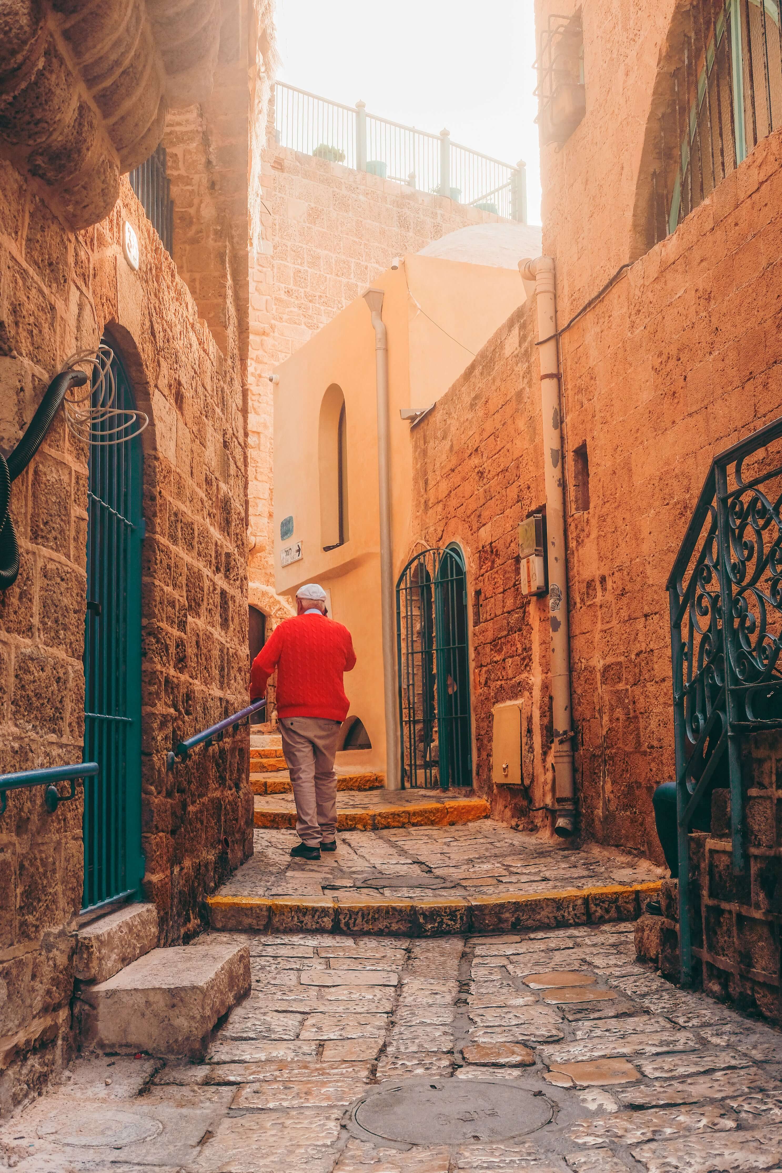 Person in a red jacket walking down a narrow stone alleyway in an old town.
