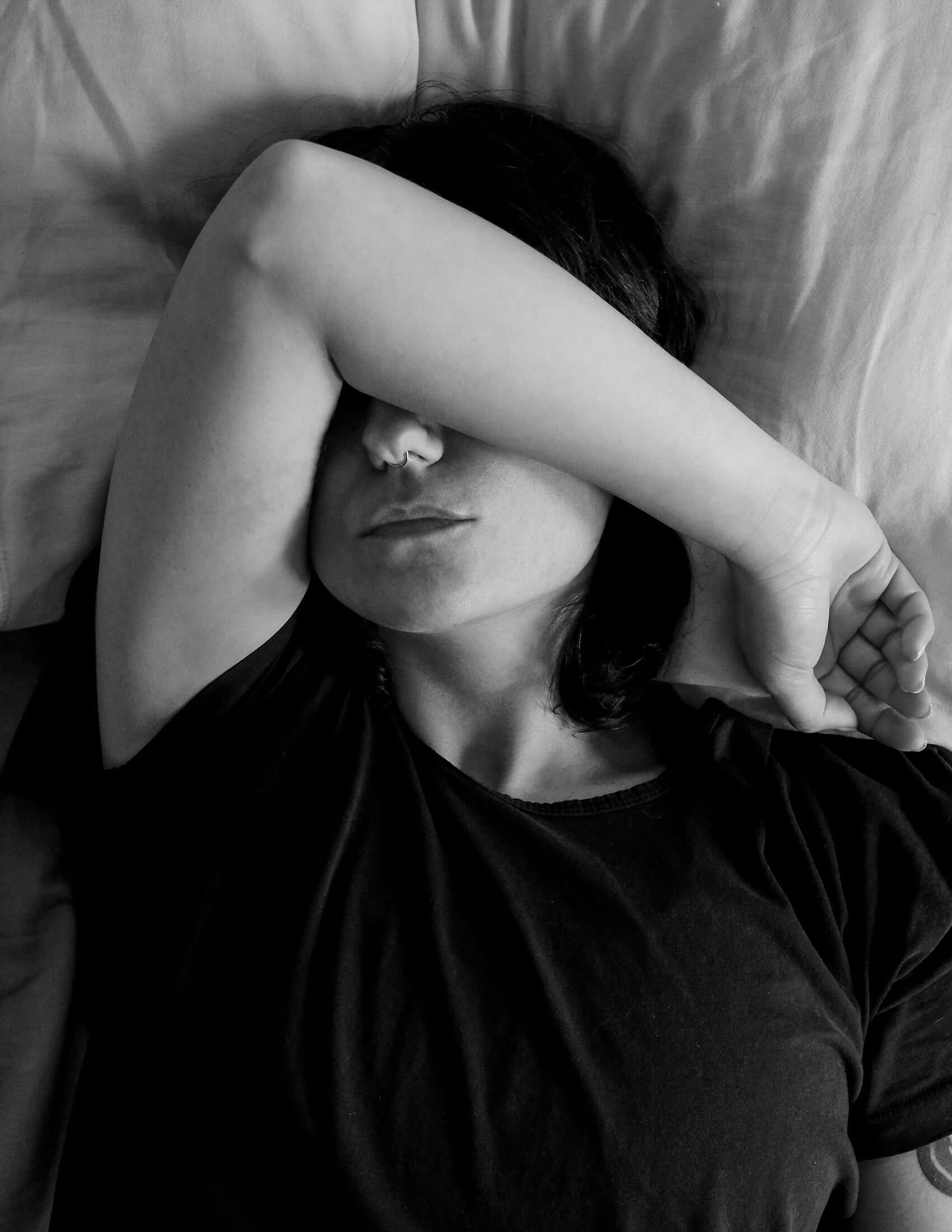 Person lying on a bed with one arm covering their face, black and white photo

