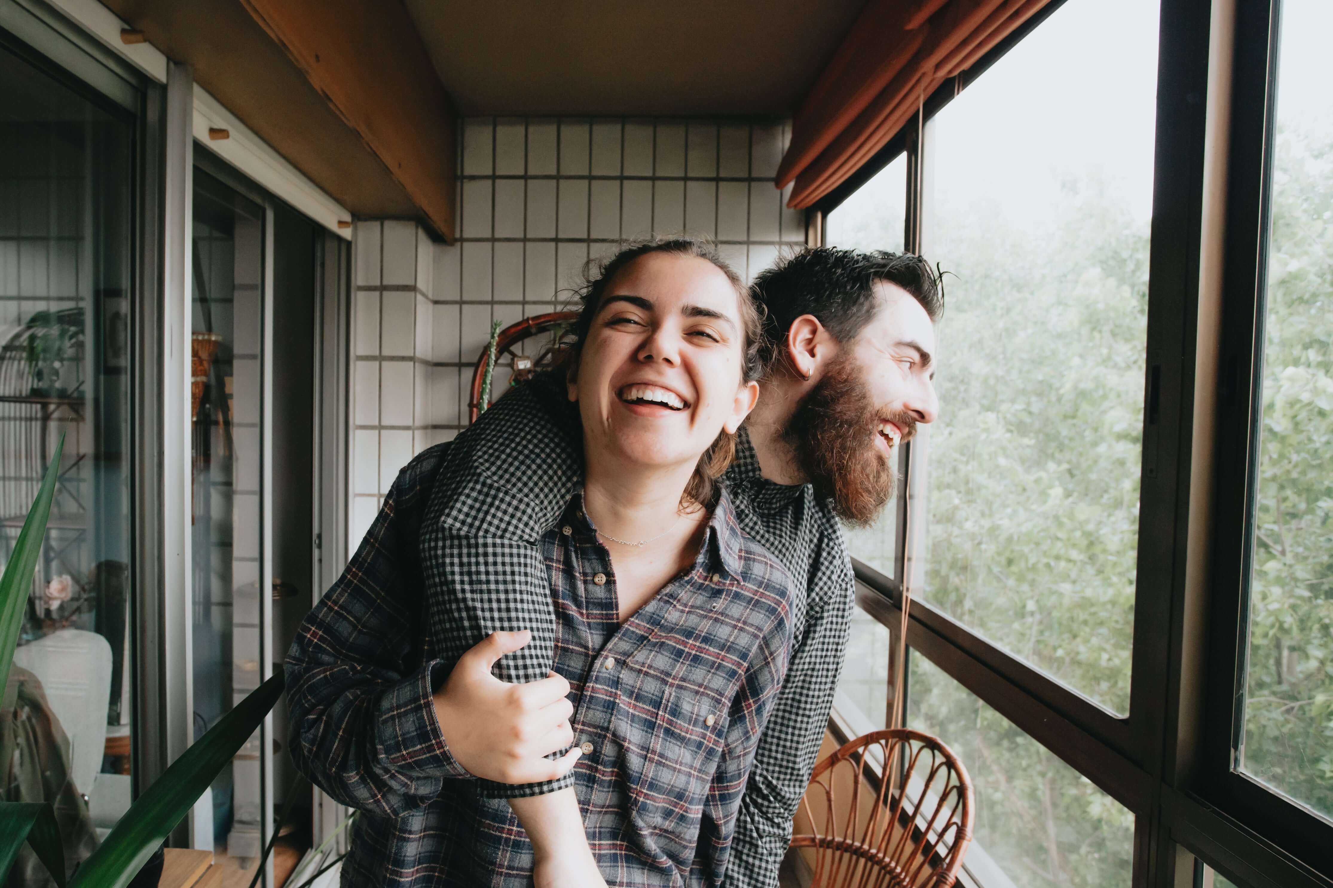 Two people laughing together on a balcony with a view of trees.
