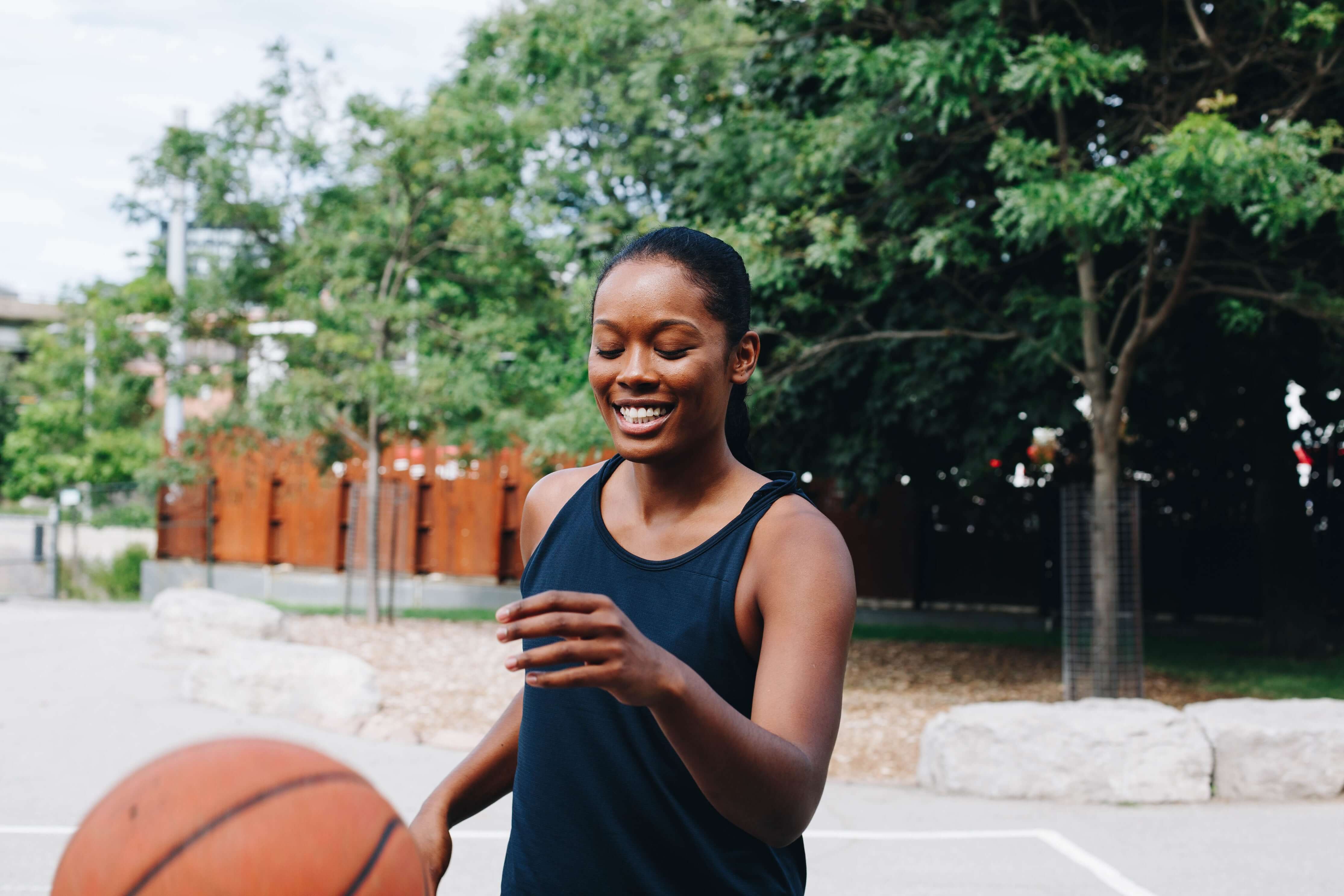 Woman holding a basketball on an outdoor court with trees in the background
