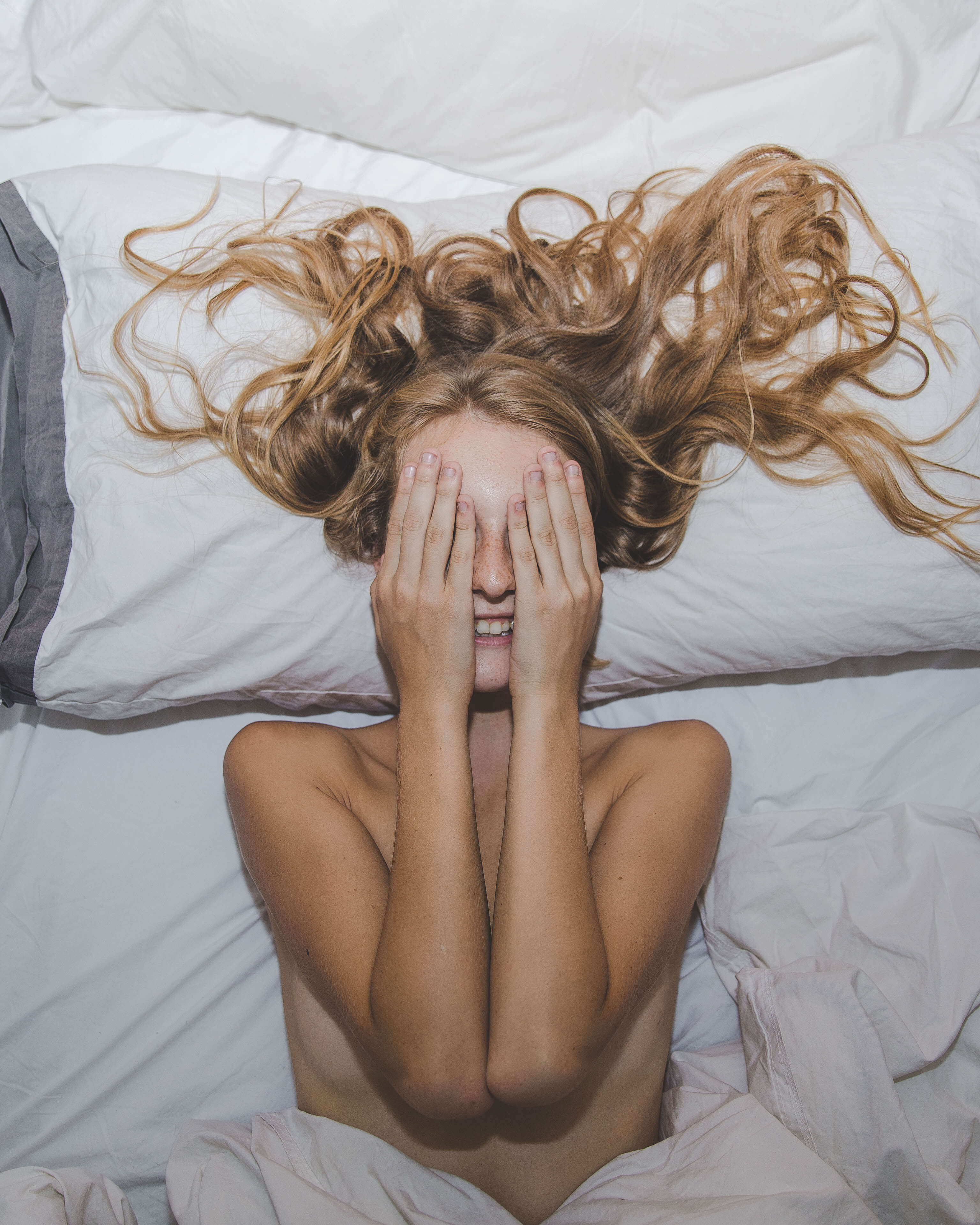 Naked person lying on a bed with long hair, partially covered by white bedding.
