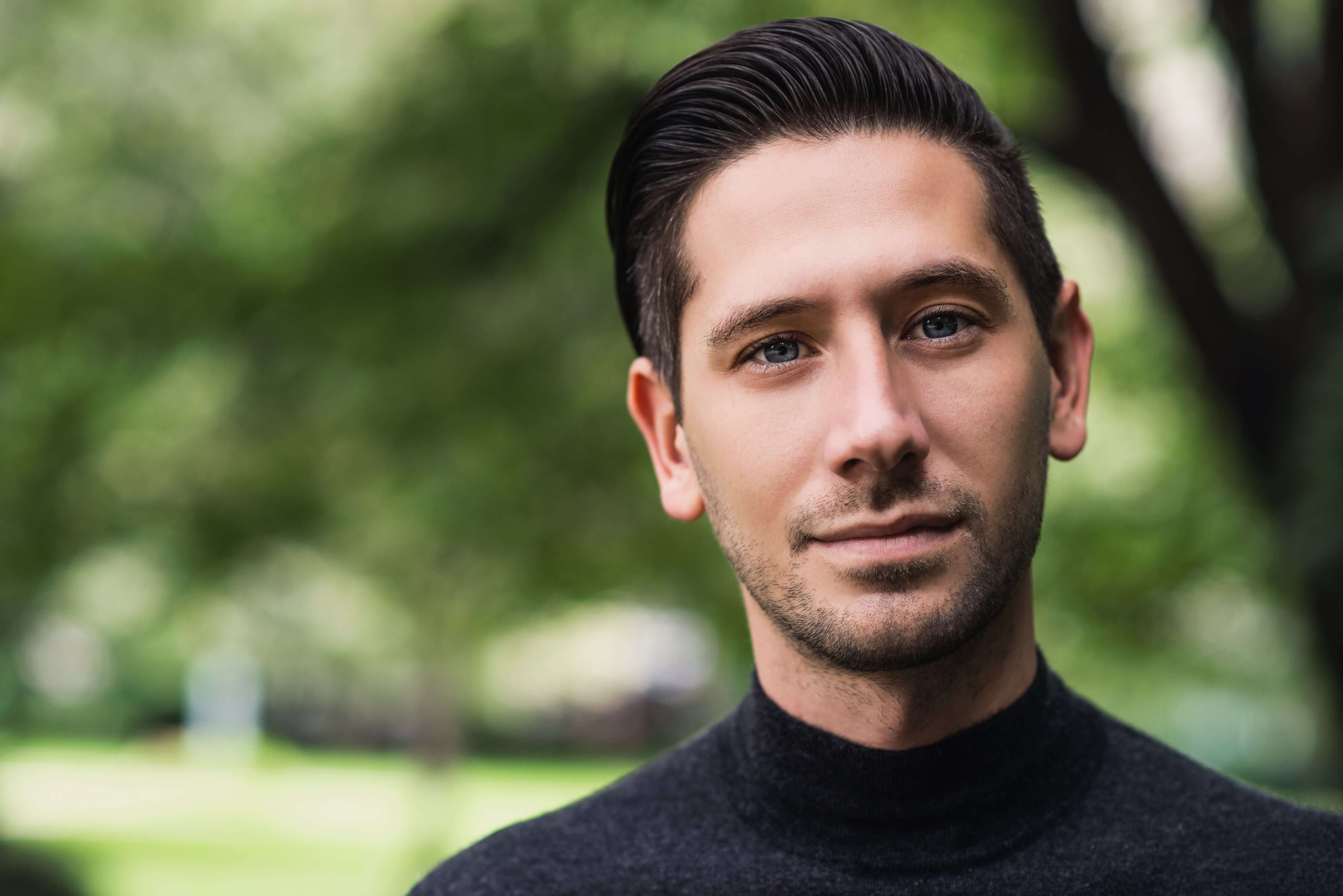 Man wearing a black turtleneck sweater with a blurred green outdoor background
